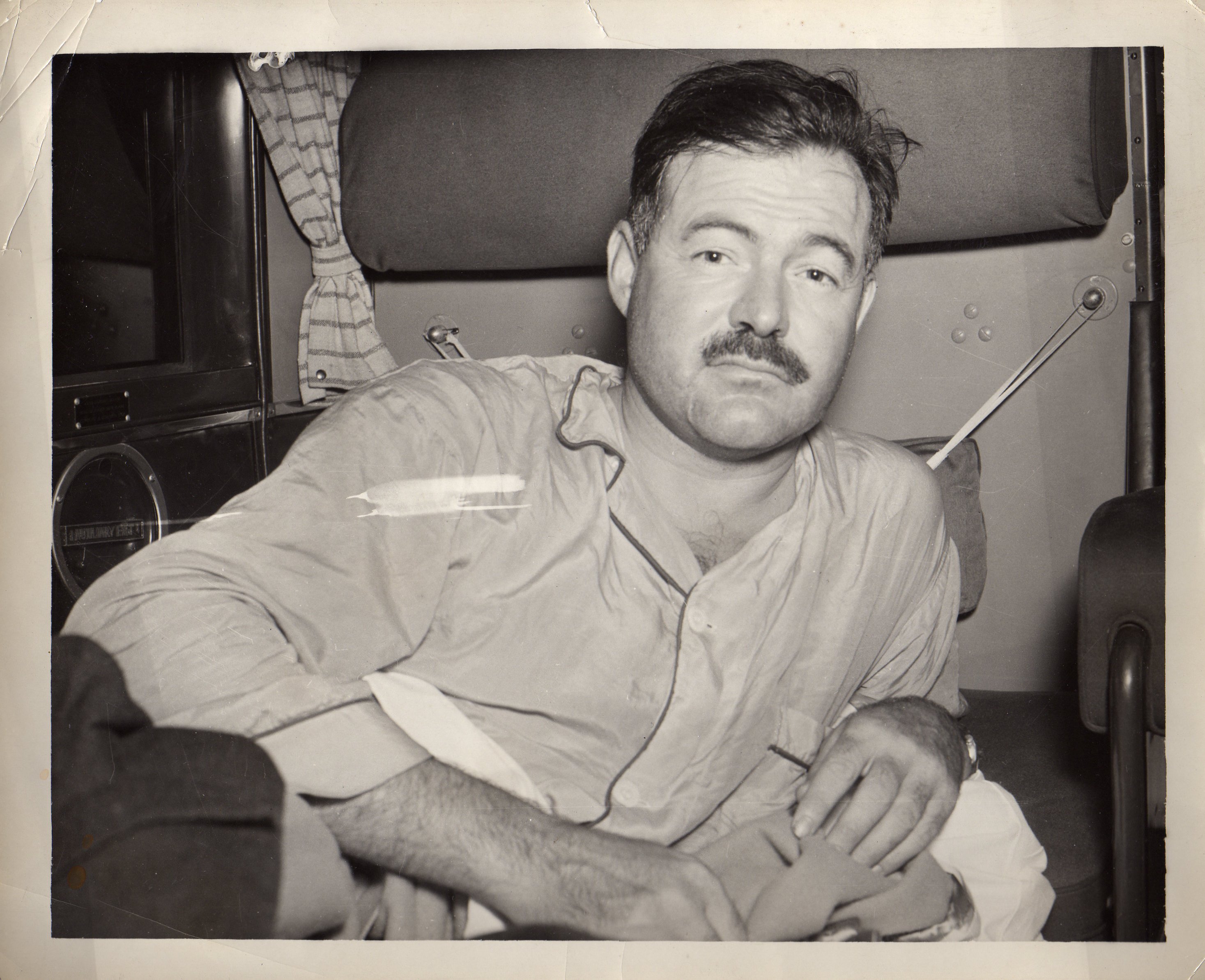 A black and white picture of Hemingway in his pajamas, just awakened, in his compartment on a sleeper plane.  He is shown from the chest up, semi-reclining, leaning on an elbow, holding the blanket in his hands.  His eyebrows are slightly raised, and he has a bit of a dazed expression.