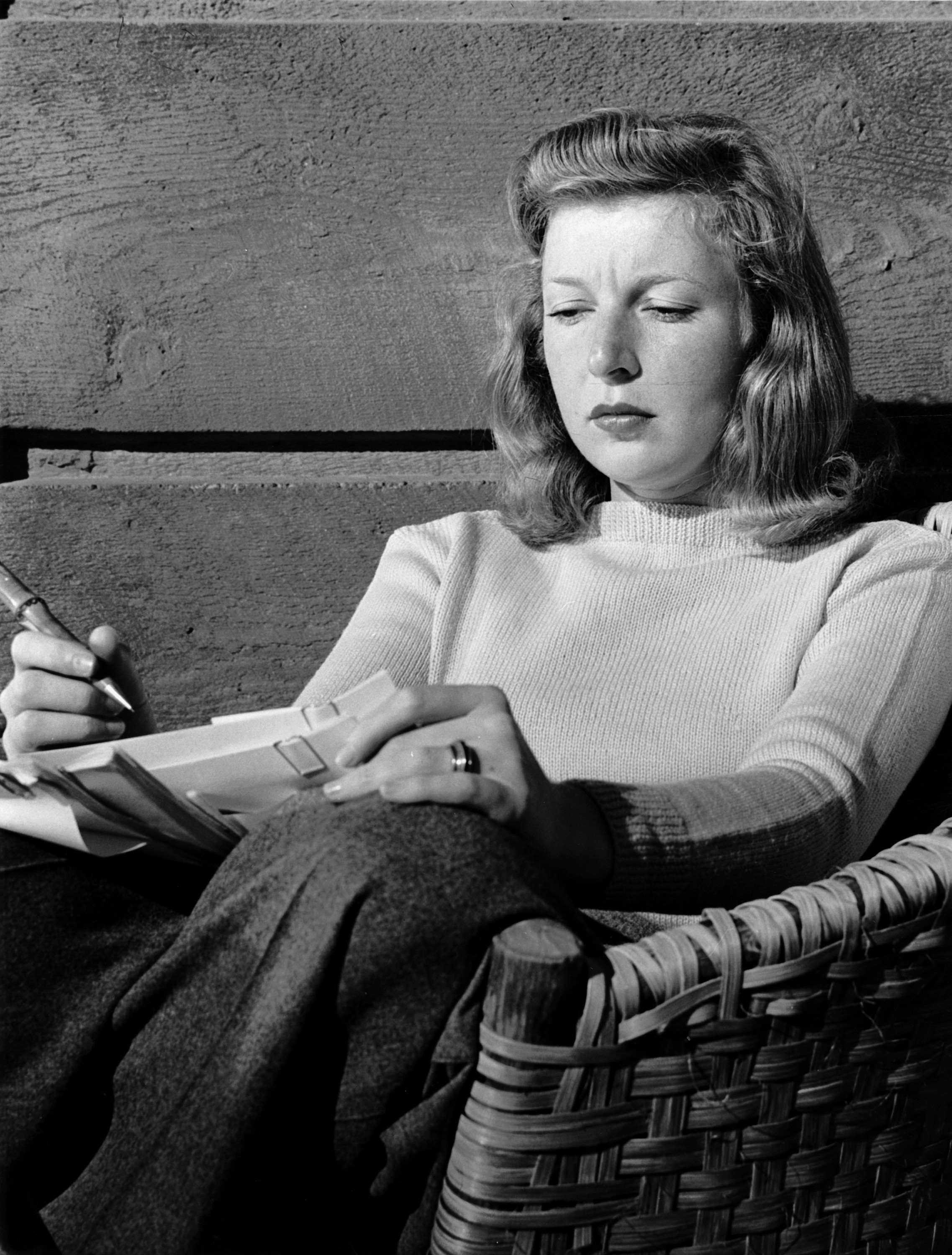 A black and white image of Martha Gellhorn writing.  She sits on a rustic chair before a wall of raw boards.  She holds papers and a pen.  She is wearing a knitted turtleneck and tweed pants, and sports a wedding ring.