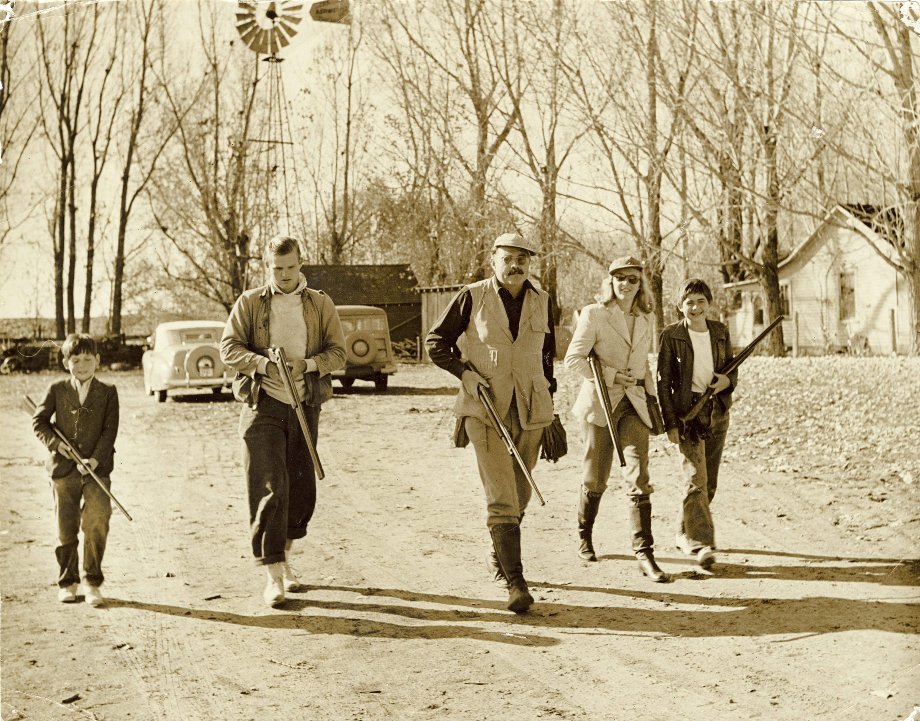 A sepia image of Gregory, Jack, Ernest, and Patrick Hemingway with Martha Gellhorn.  They are all carrying rifles and are dressed for hunting.  They walk toward the camera; behind them two parked cars, an outbuilding, and a white house are visible.  Behind those are a line of trees marking the edge of a field.
