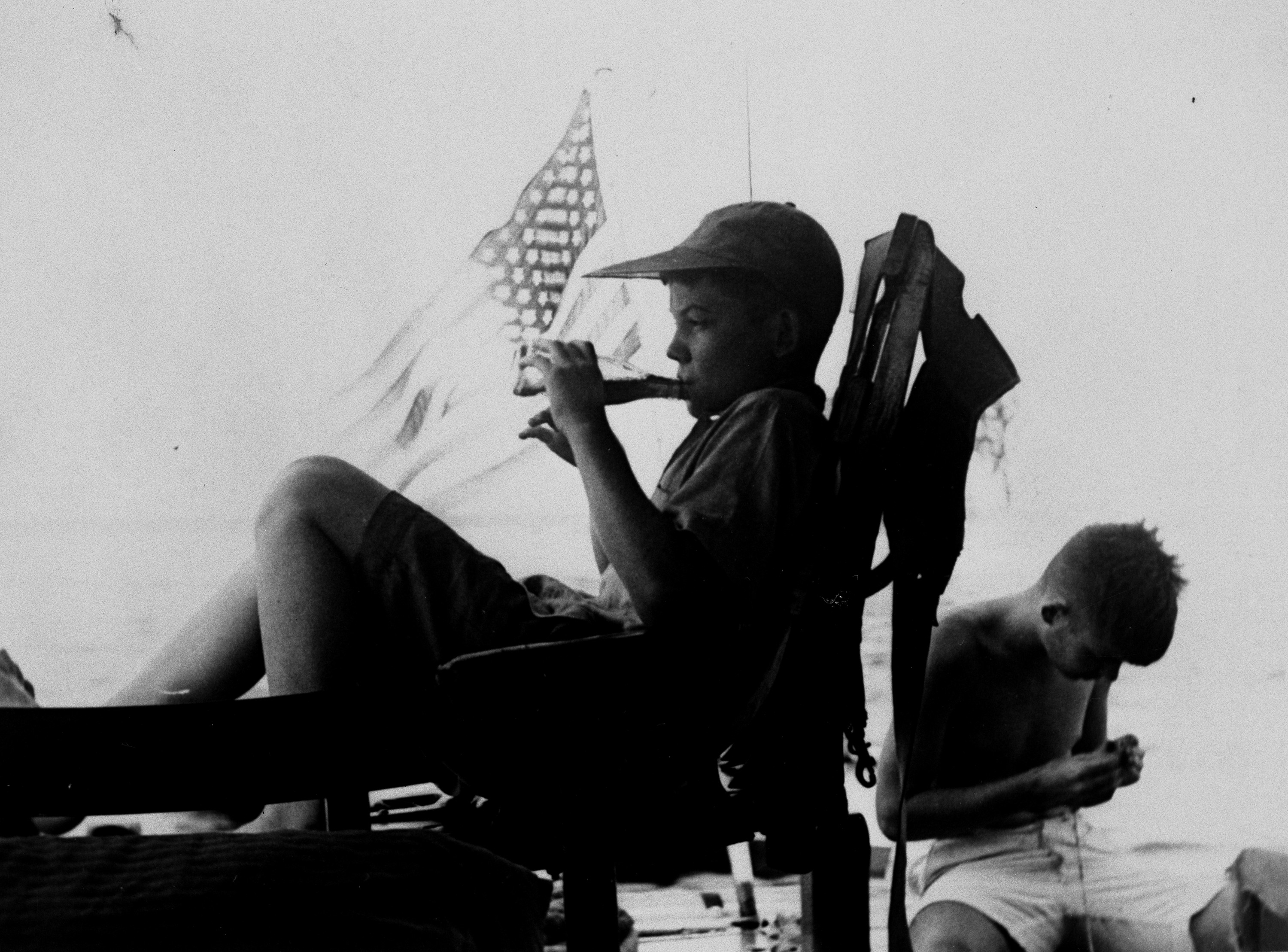 A black and white photograph of Hemingway's teen-aged children Gregory and Patrick aboard Hemingway's boat.  Gregory, seated in the foreground, wears a billed cap and is drinking from a clear glass bottle.  Patrick, in the background, wears white shorts and is concentrating on something small he holds in his hand.  The United States flag flies as ensign off the stern of the boat.