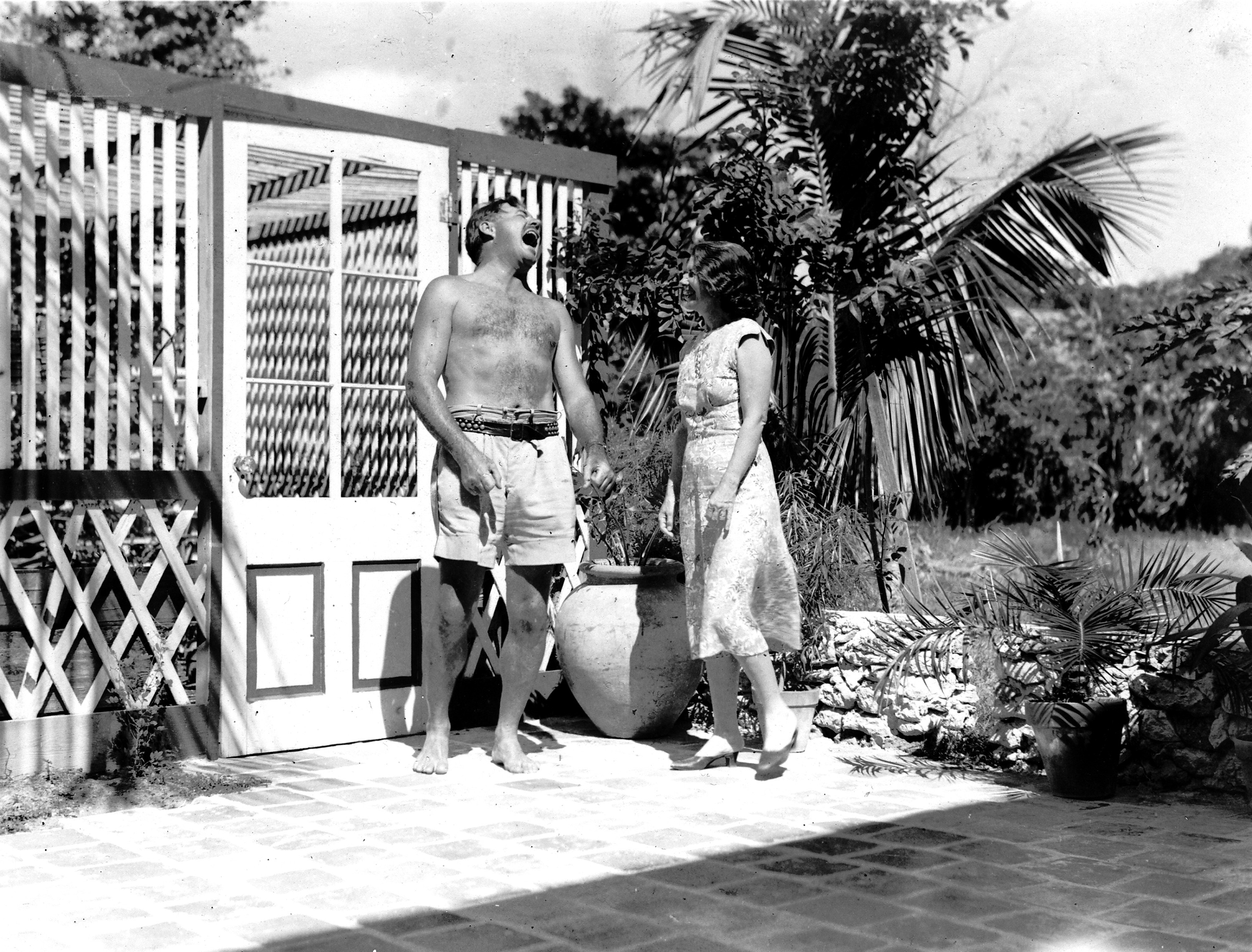 A black and white image of Ernest and Pauline Hemingway laughing by their home in Key West.  Ernest wears only shorts and a belt; Pauline wears a sleeveless dress and flat shoes.  Ernest's head is thrown back in laughter, Pauline laughs, looking at him.  They stand on a tiled patio next to a palm tree.