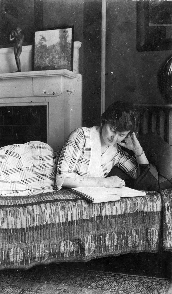A black and white photograph of Hadley Hemingway lying on a bed, reading.  The bedspread is an abstract, multi-hued woven material.  Hadley leans her head on her legt hand.  She is wearing a checked summer dress, mostly light-colored, with darker checks, a white collar, and a pocket.  Behind her is a fireplace; on the mantel are a small square landscape painting and a statue of a nude woman holding a water vessel.  Barely visible behind her to the right is a steam radiator.