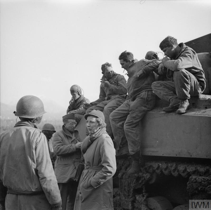 A black and white image of Martha Gellhorn standing next to a tank, talking to soldiers.