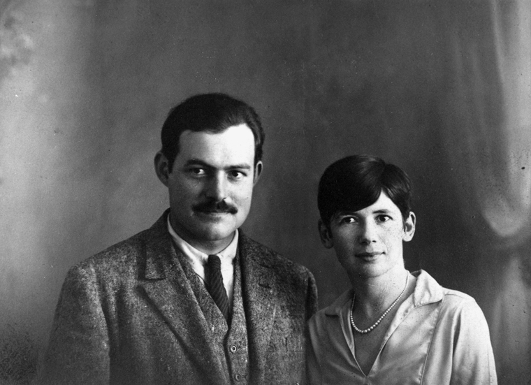 A black and white formal portrait of Ernest and Pauline Pfeiffer Hemingway standing before a photographer's background.  Ernest wears a tweed jacket, vest, white shirt, and tie; Pauline wears a white silky blouse and a strand of pearls.  Both have dark hair and look seriously at the camera.