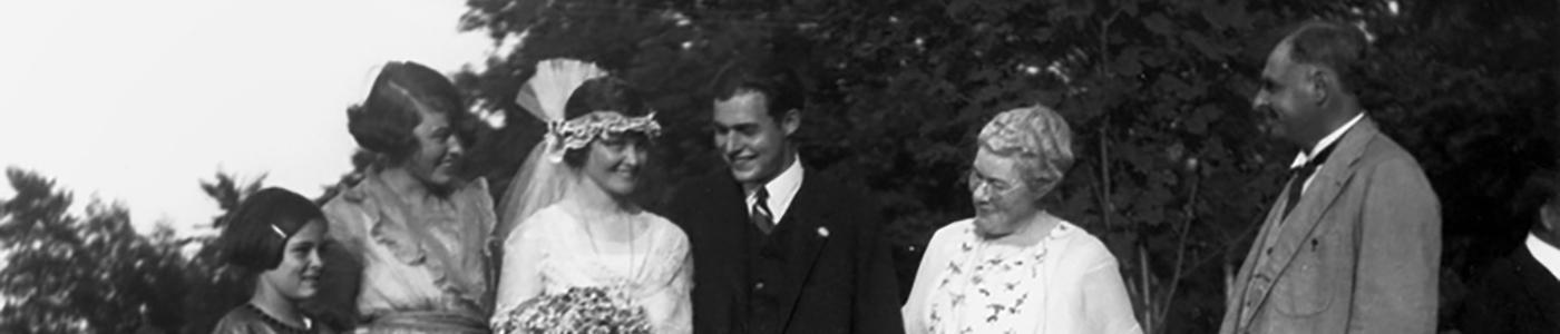Hadley and Ernest Hemingway smiling on their wedding day, surrounded by Hemingway's parents and sisters.  Hadley wears a white wedding dress, low headband, and veil; Ernest is in a white shirt with a dark tie, vest, and blazer. The photo was taken outside against a backdrop of trees.
