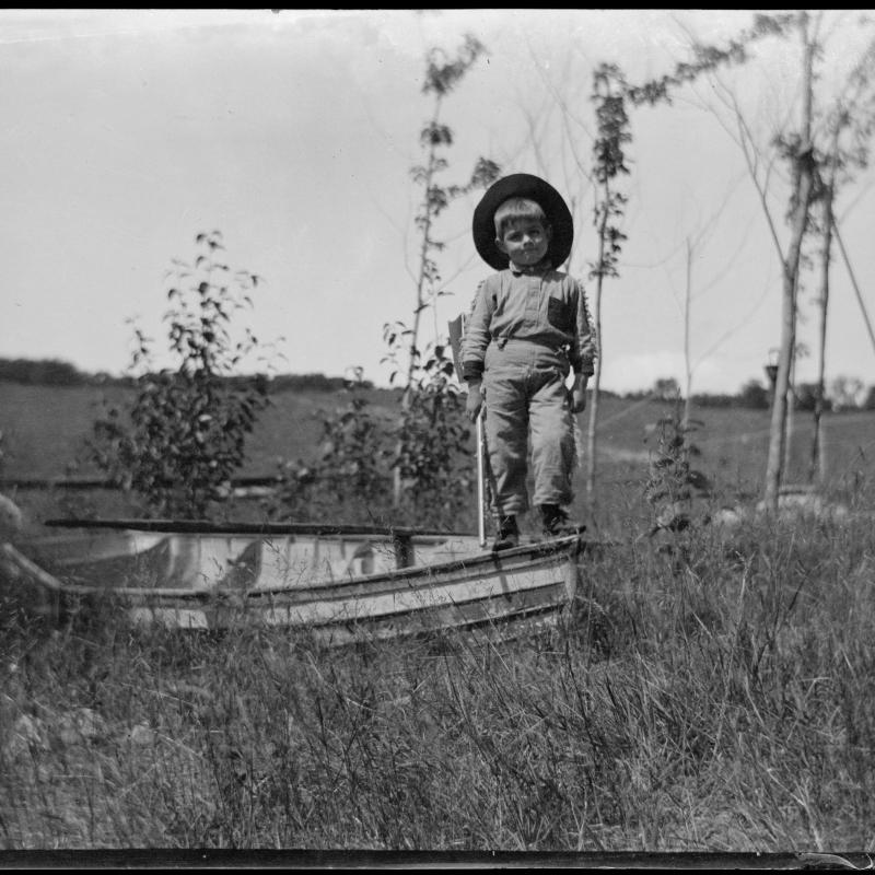 Hemingway, age 4, stands on the prow of a boat that sits in a field.  He is wearing a hat and a fringed Western costume, and he holds a toy rifle.