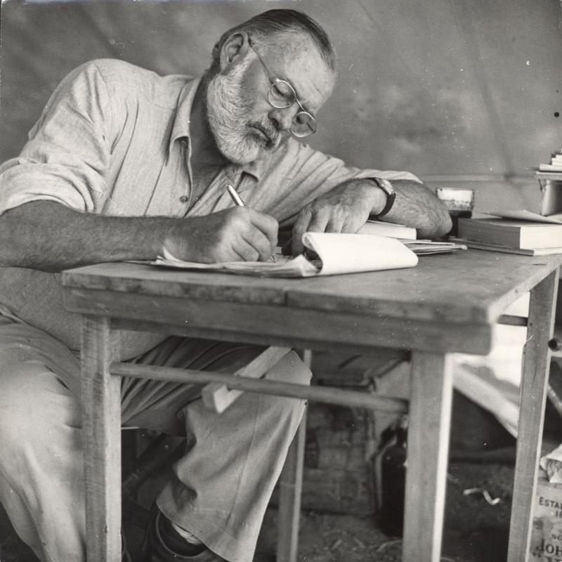 Hemingway writing at a camp table in Africa, 1953-54
