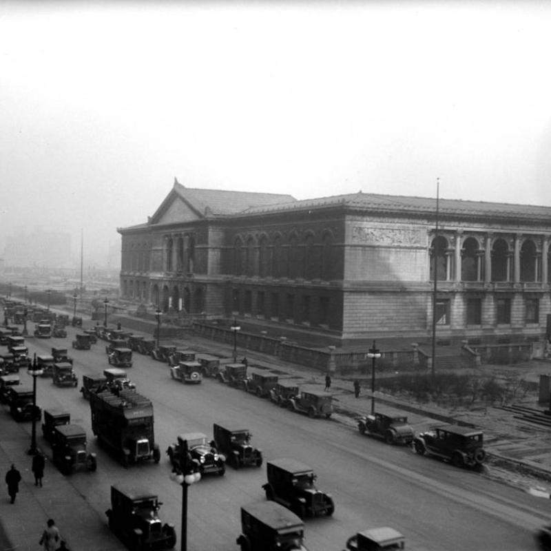 A black and white image of the Art Institute of Chicago, street view, taken 1920s. Cars and trucks line the street, and traffic flows in two directions. Several people walk down the near sidewalk.  