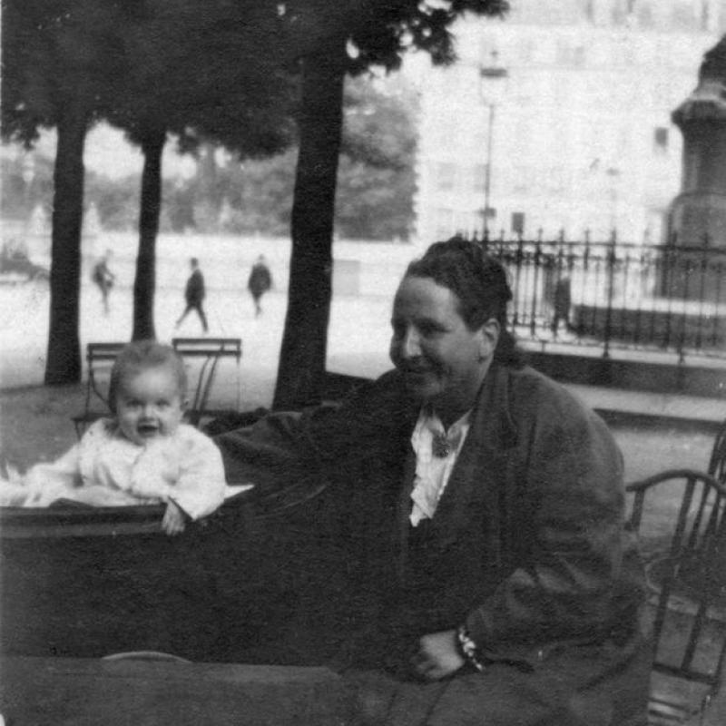 A black and white image of Gertrude Stein with a baby Jack (Bumby) Hemingway in his pram.  Taken in a park in Paris, late 1920s.  Stein, wearing a dark colored jacket and skirt with a white blouse under the jacket, and baby John are both smiling.  Stein has her arm around the baby; she is seated on a metal chair next to his pram.