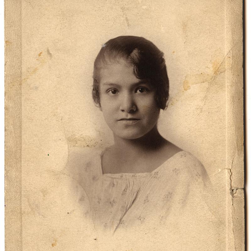 Photograph of a studio portrait print of a young Anishanaabe girl wearing a light, pale flower-patterned blouse.  Her hair is pulled back save for a waved bang over her left eye. Shown from the chest up, she has brown eyes and gazes directly at the camera.  Embossed into the card stock is the studio name, Friest, and the location, Charlevoix. 