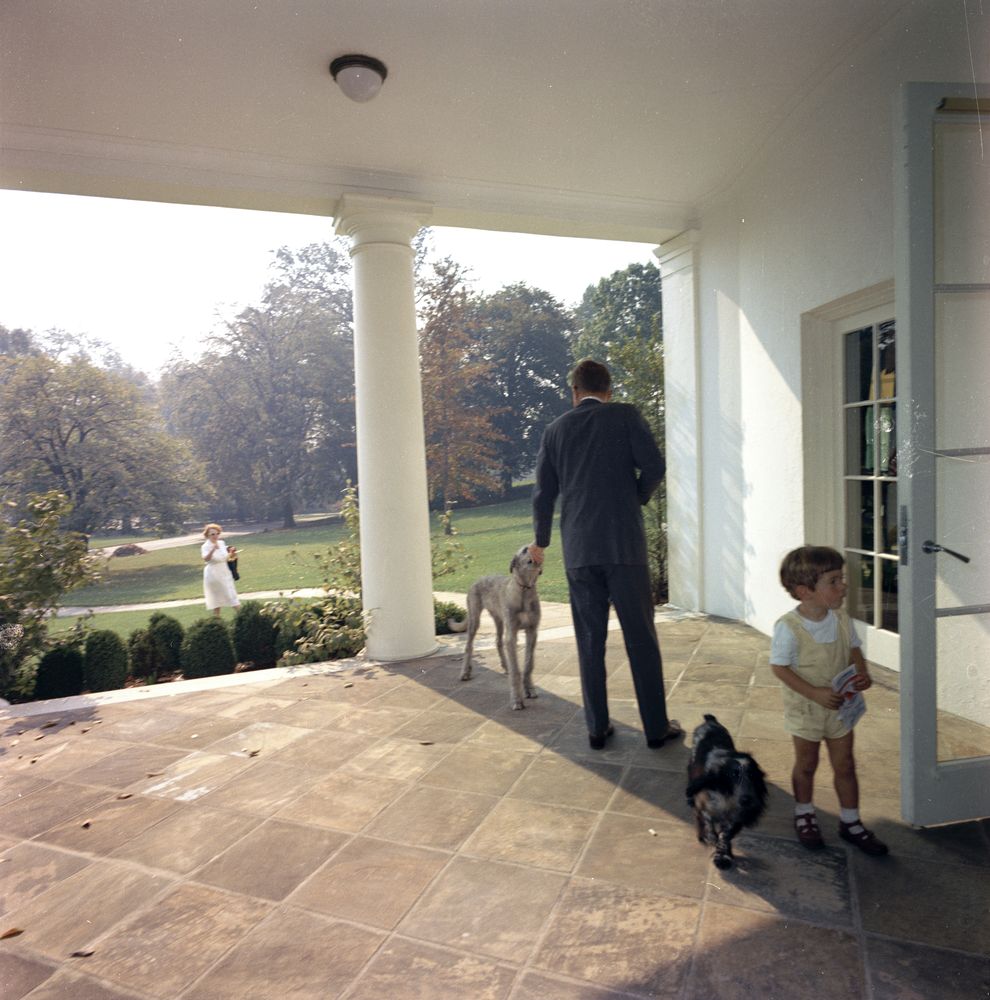 JFK feeds his dog, Wolf, on the West Wing Colonnade of the White House, Washington, D.C. John Jr. (right), stands with family dog Shannon near open door to the Oval Office; nanny to the Kennedy children, Maud Shaw (at left, in background), looks on.