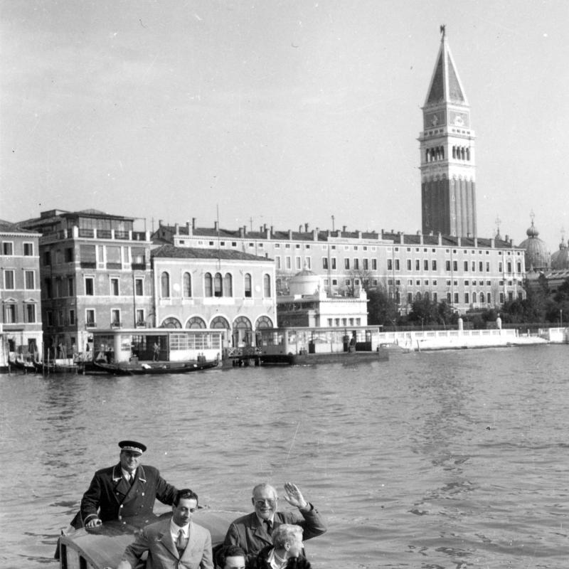 A black and white photograph of Ernest and Mary Hemingway in a launch in Venice.  Hemingway's hand is raised in greeting.  The buildings of the main island (with the St. Mark's campanile) in the background.